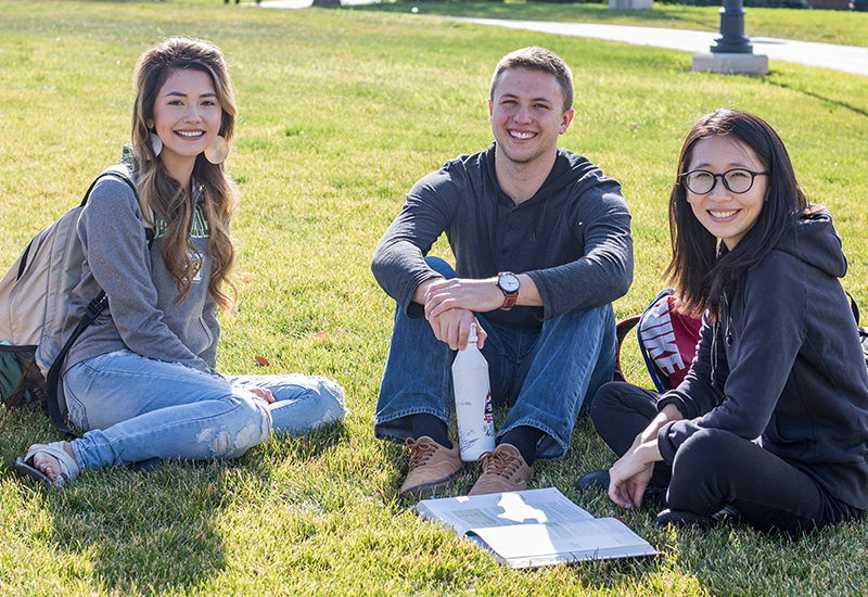 Three students sit on the campus green and pose for a photo.
