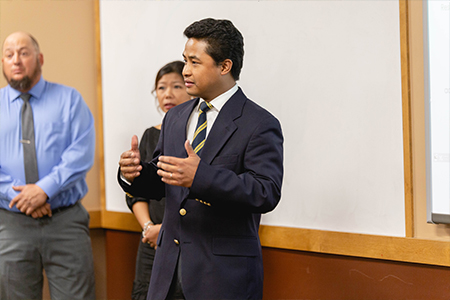 A man gives a presentation with two people in the background.